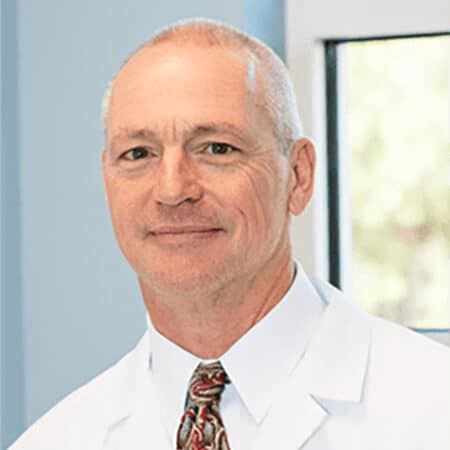 Man in white lab coat smiles at the camera, tie visible, light setting.