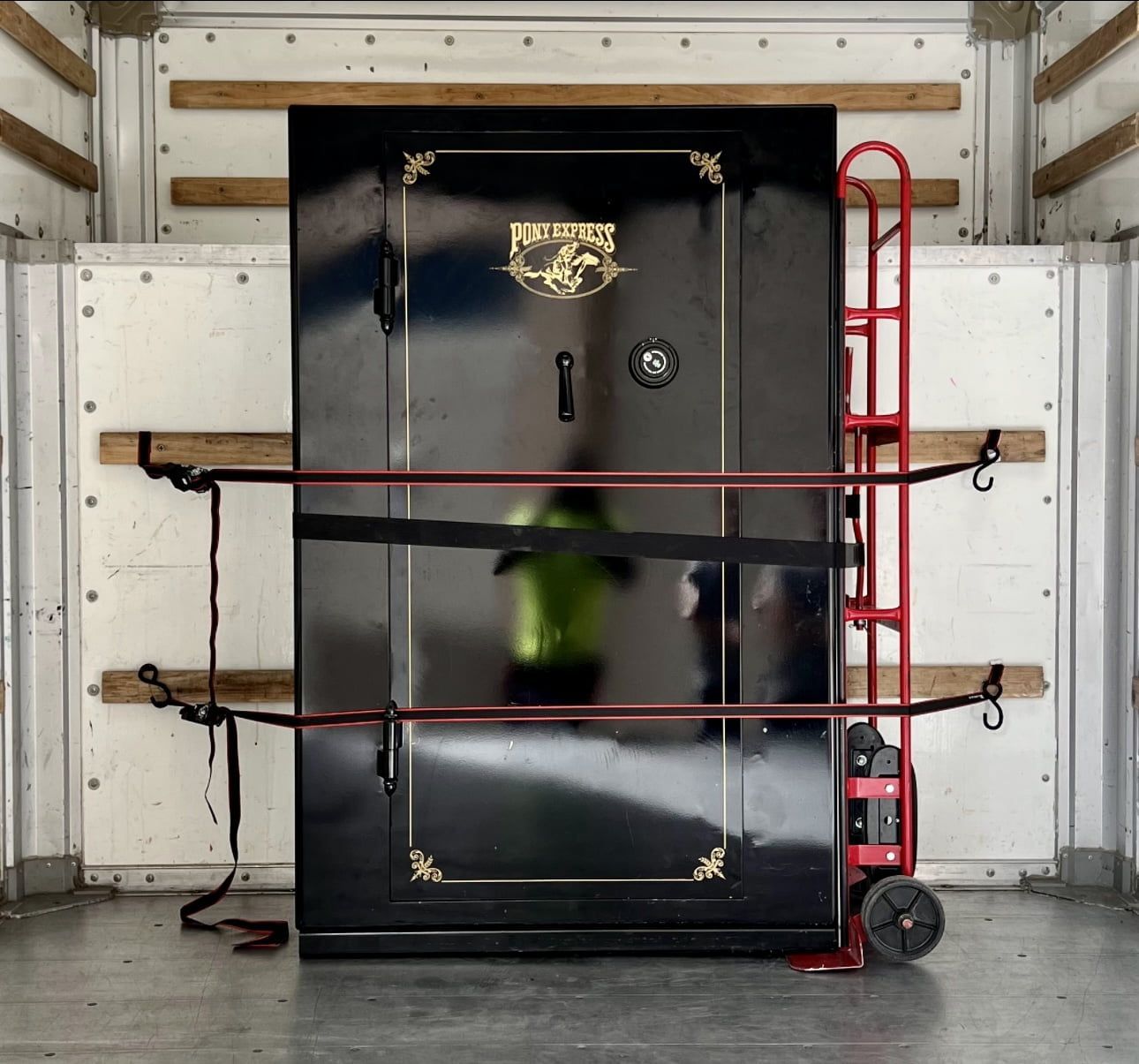 A black gun safe secured with straps inside a truck, with a red hand truck beside it.