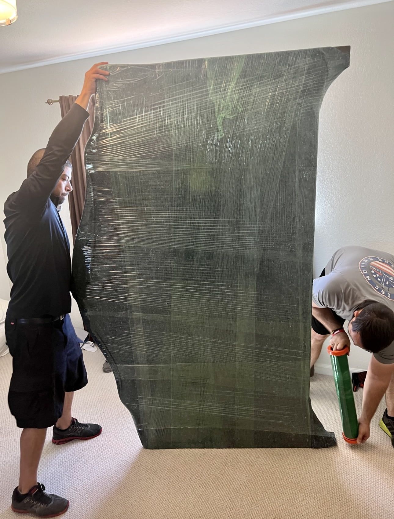 Two people holding and moving a large, wrapped, dark-colored object indoors on carpet.