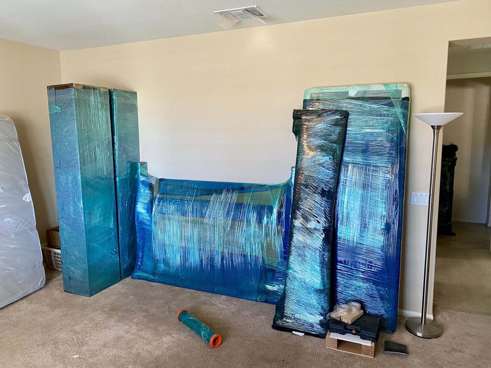 Large abstract blue and turquoise artworks leaning against a beige wall, a lamp, and a partially visible doorway.
