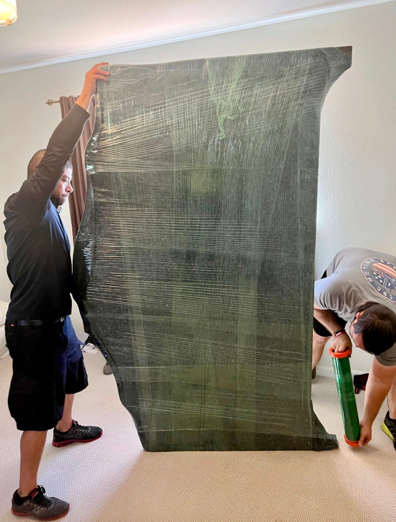 Two movers lift a gray couch in a room with boxes; one kneels, smiling, the other stands, smiling.