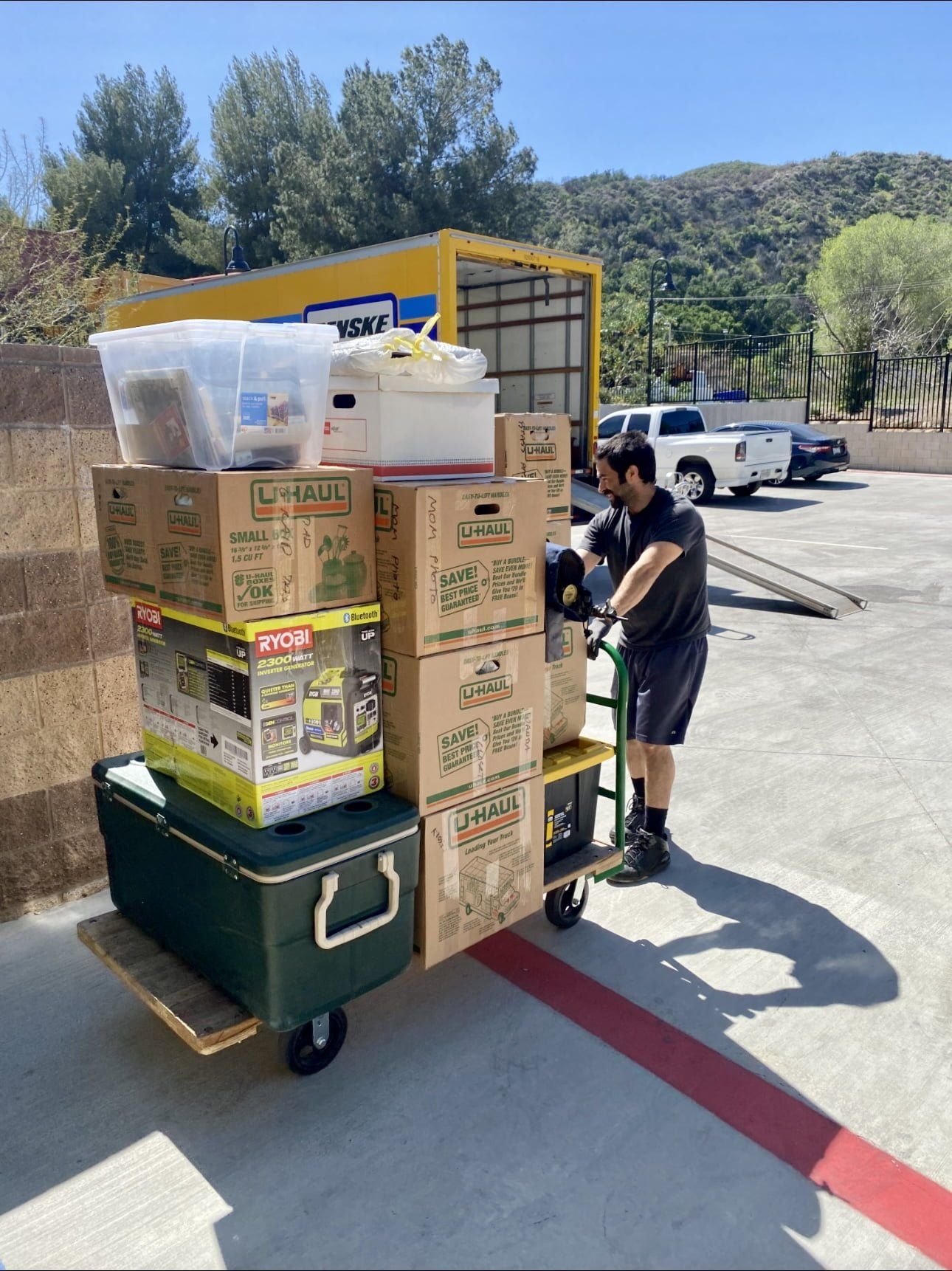 Man loads boxes onto a dolly to move into a moving truck, outdoors. Sunny day, hillside background.