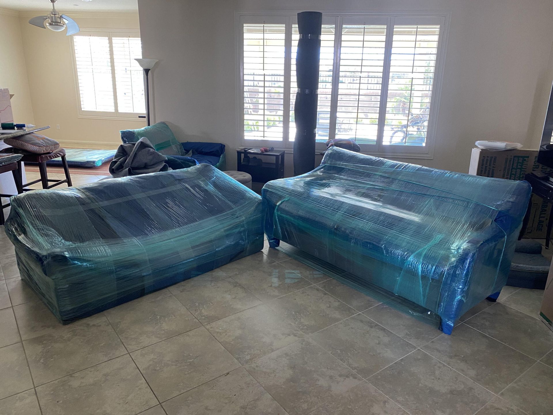 Two blue sofas wrapped in protective plastic, in a brightly lit room, possibly for moving.