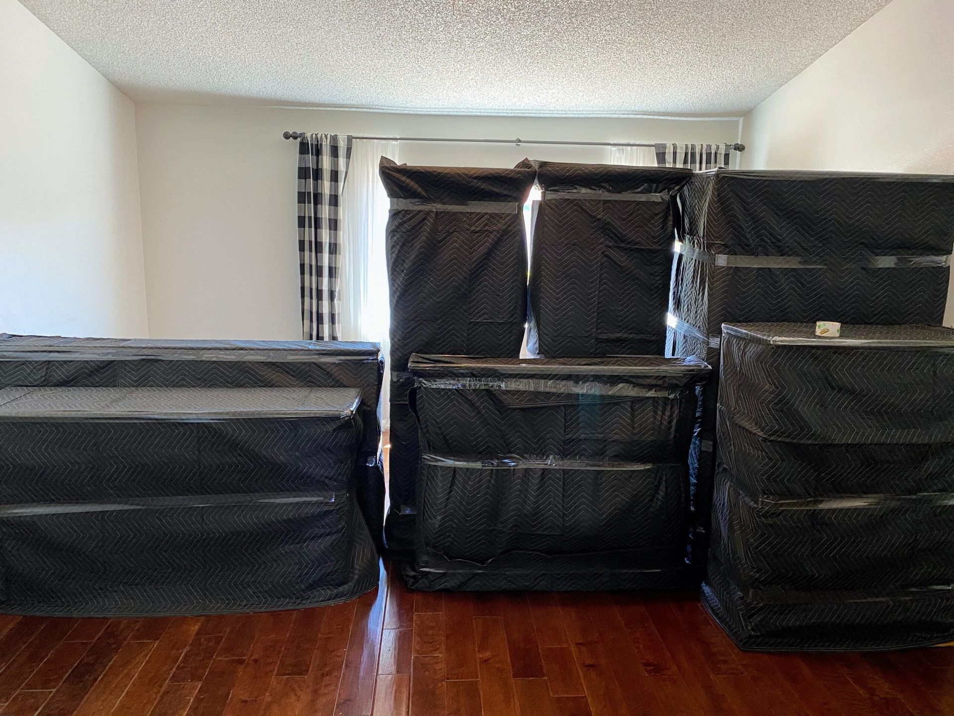 Mattresses wrapped in black plastic, stacked in a room with hardwood floors and checkered curtains.