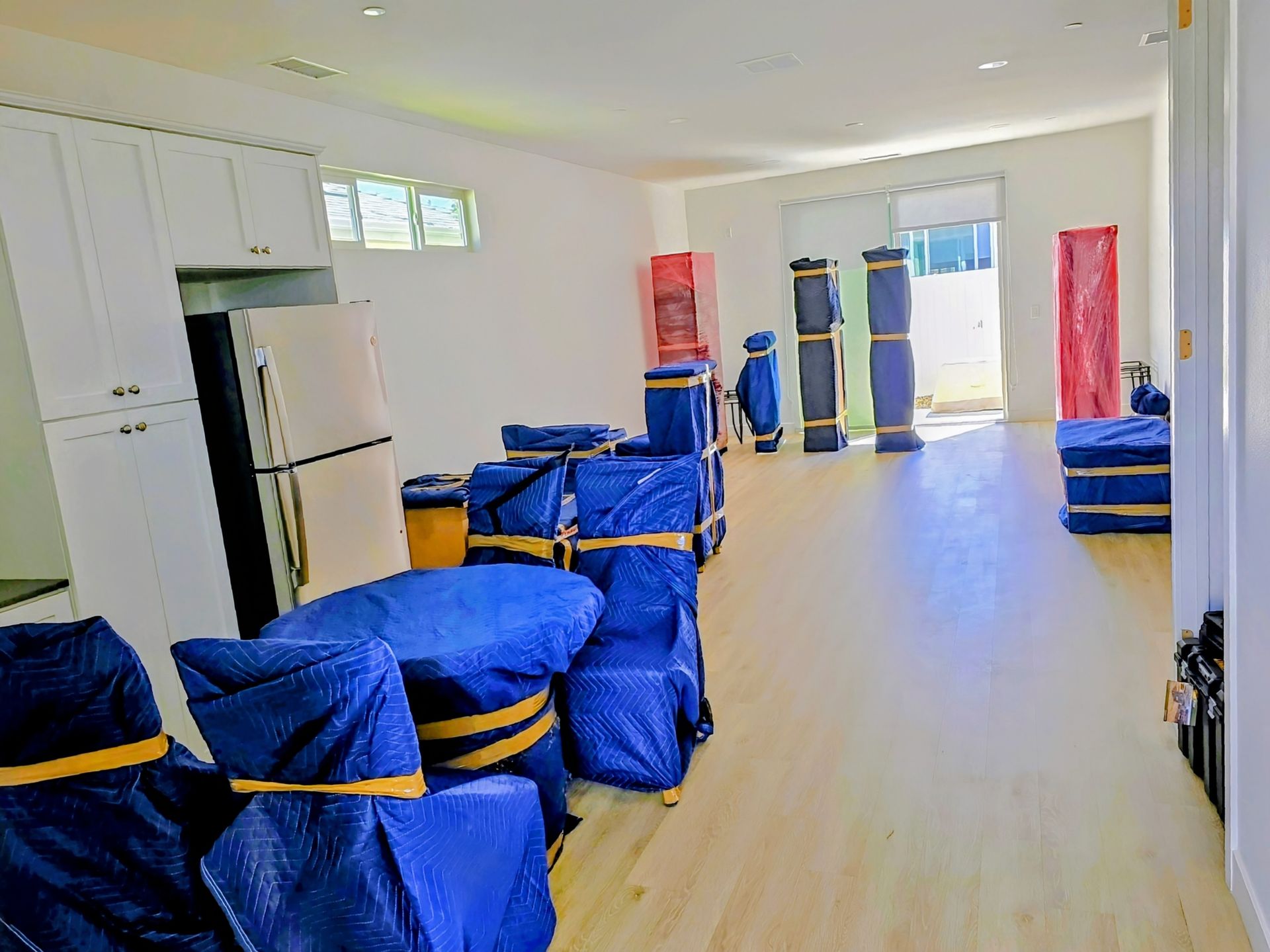 Two workers in red shirts and gray overalls move a washing machine in a kitchen.