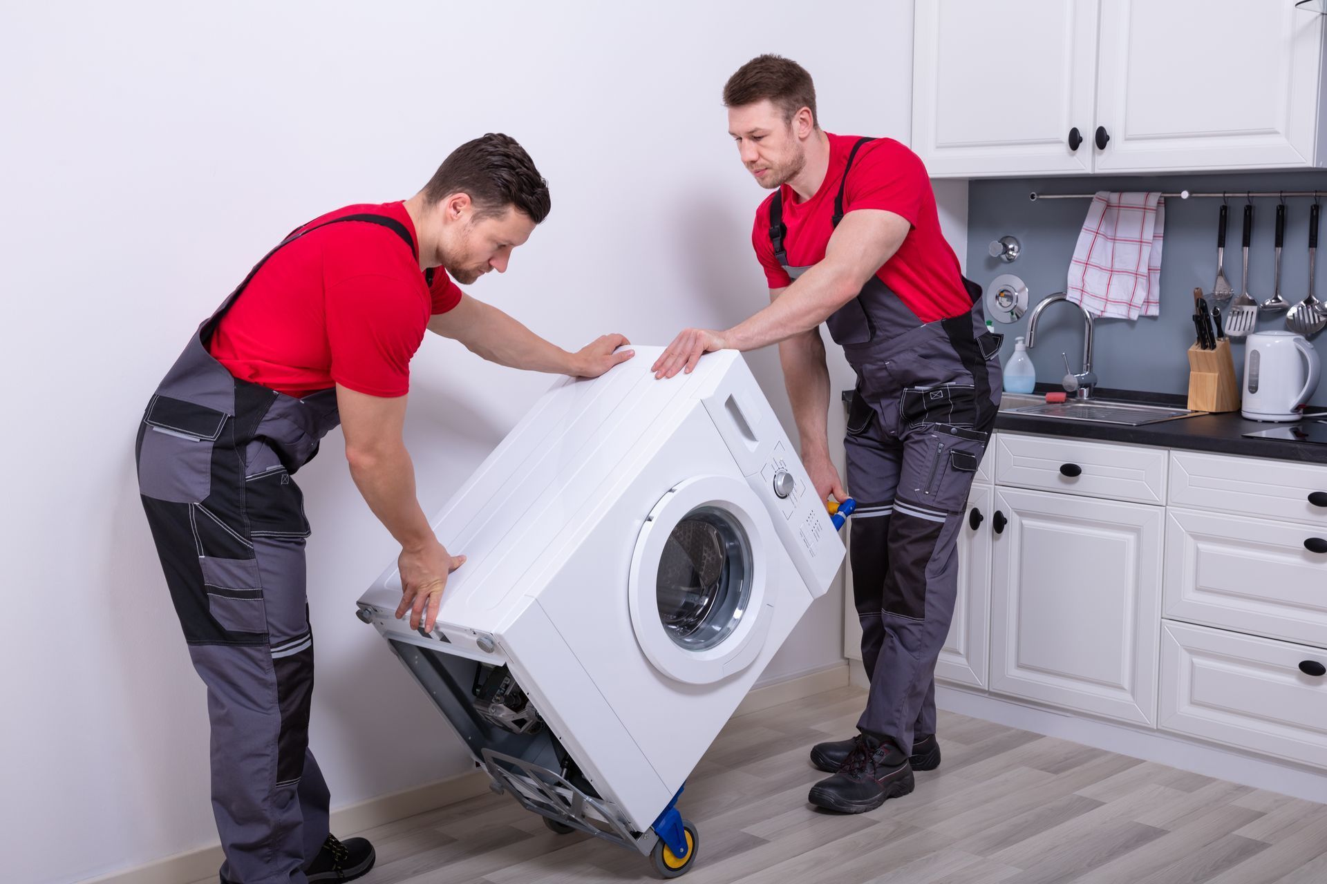 Two workers in red shirts and gray overalls move a washing machine in a kitchen.