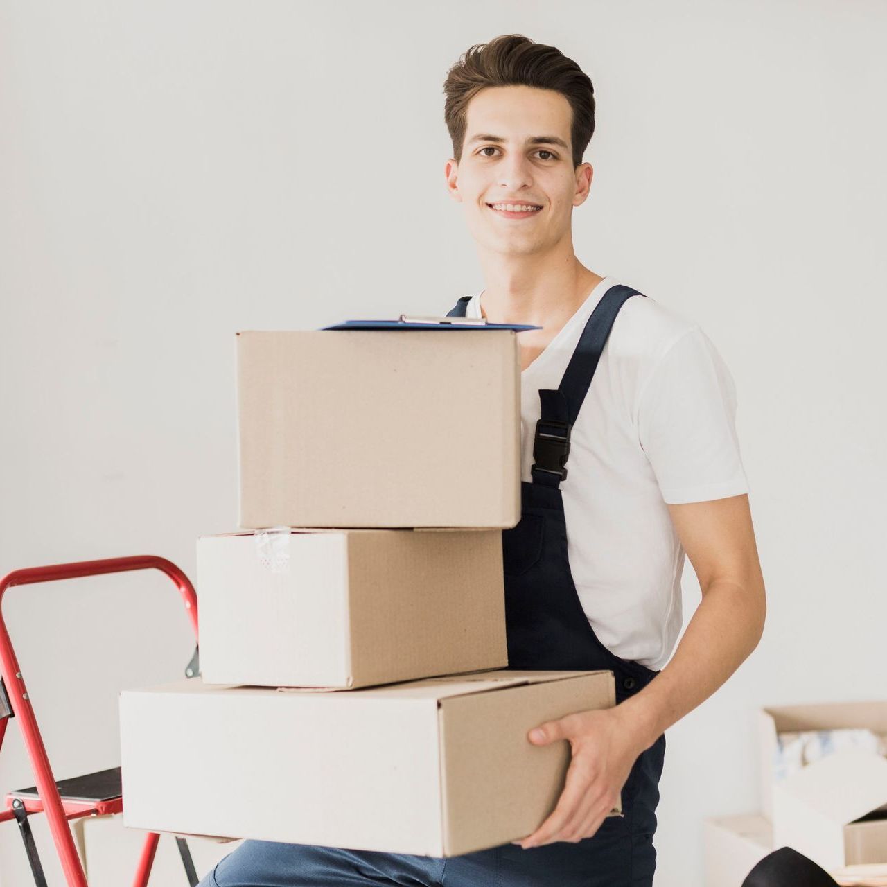 Smiling person in overalls holding three cardboard boxes. Red ladder and boxes in background.