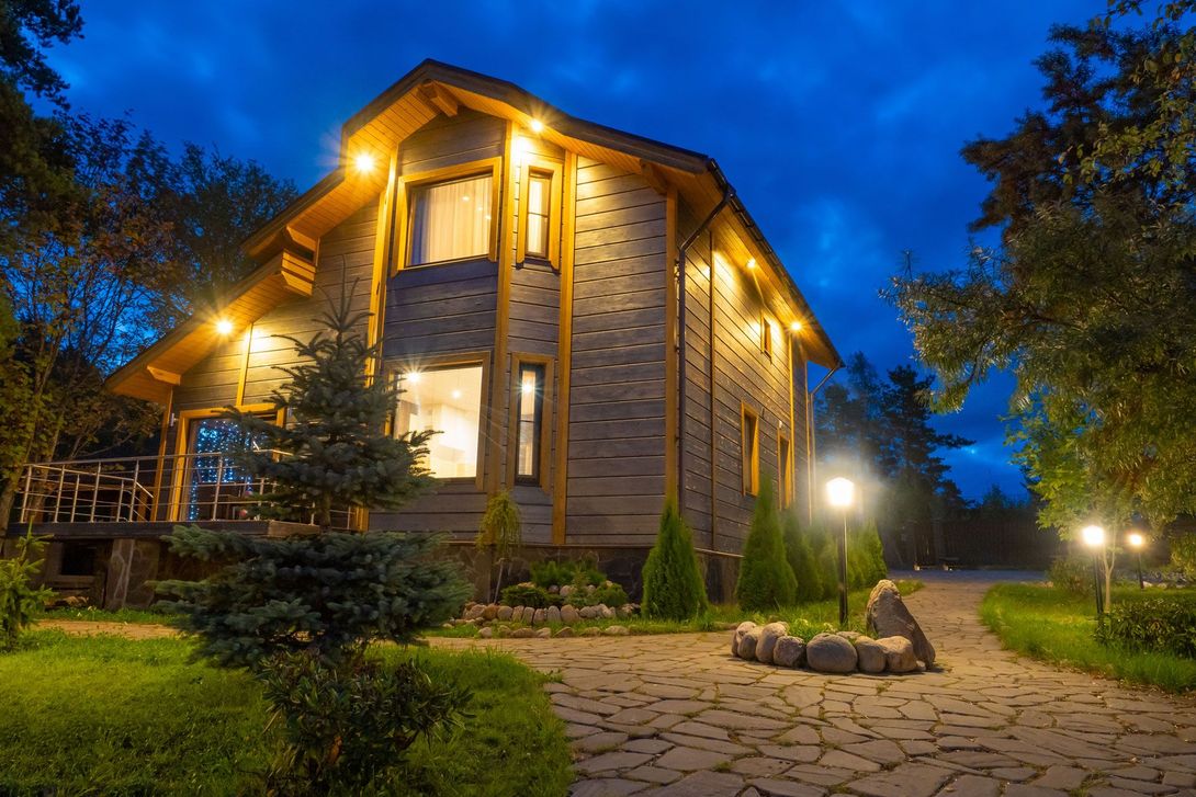 A two-story log house illuminated at twilight, set in a lush garden with a stone path and glowing outdoor lanterns.
