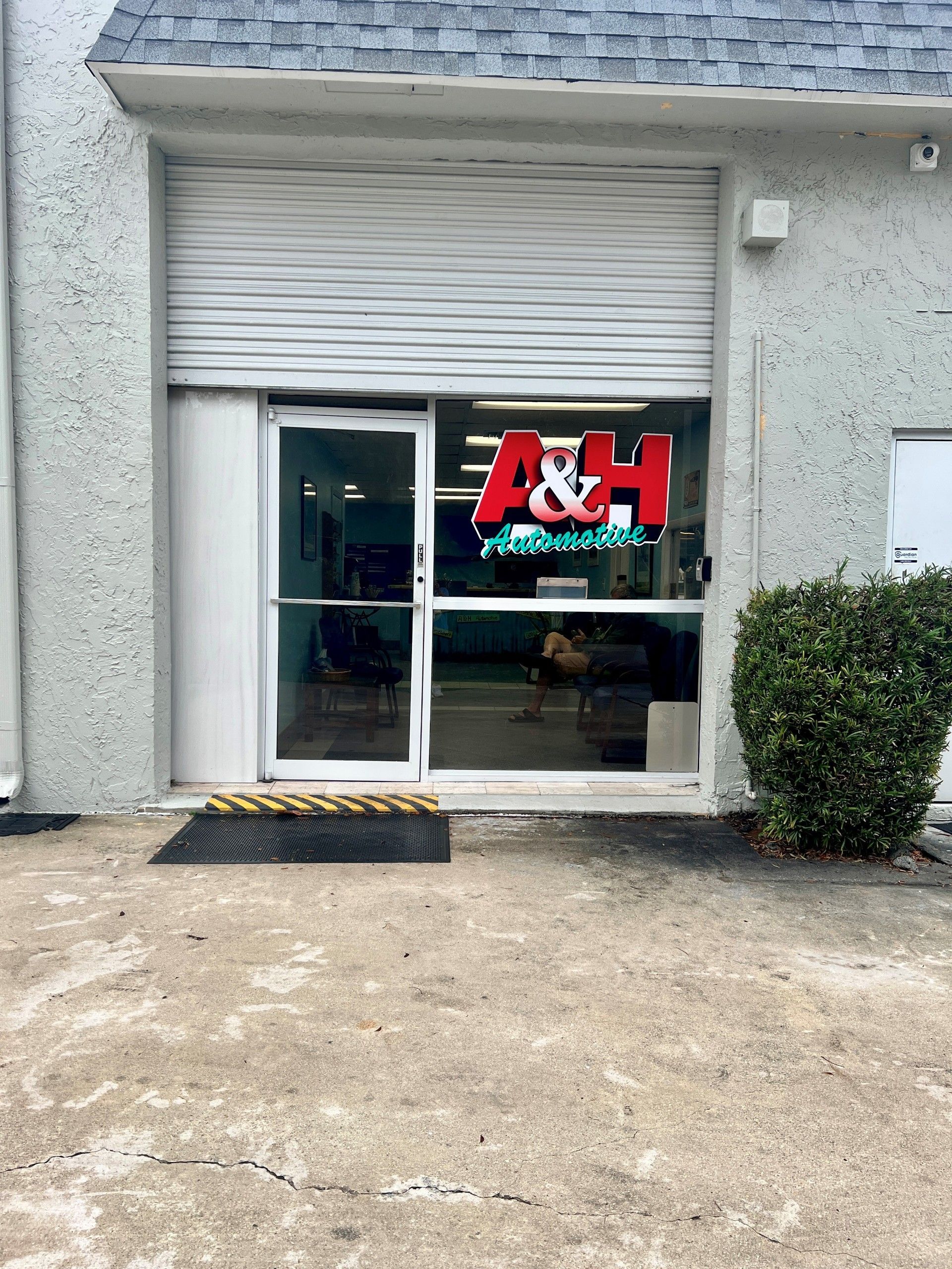 A&H storefront entrance with glass door, red sign, gray exterior, and a small bush beside the entry.