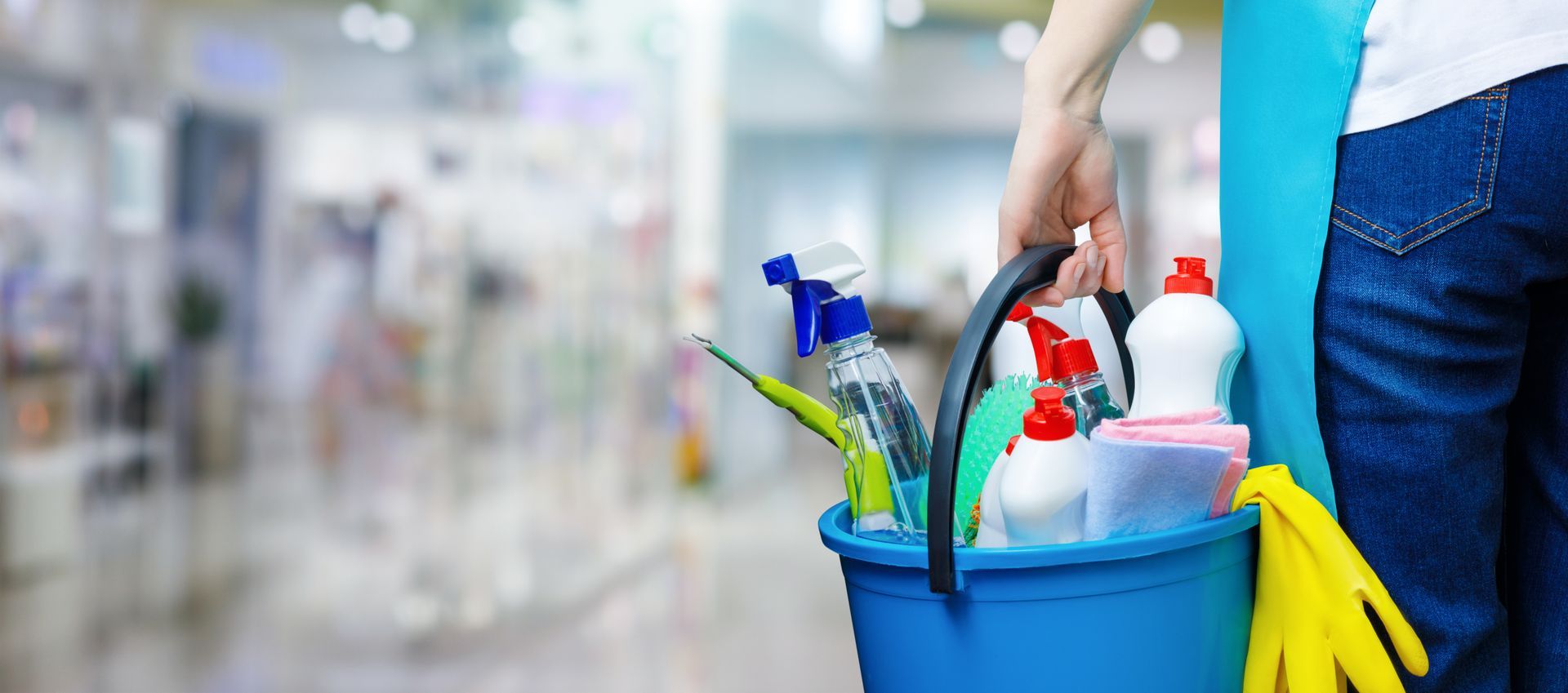 Person carrying a blue cleaning caddy with supplies in a bright living room.