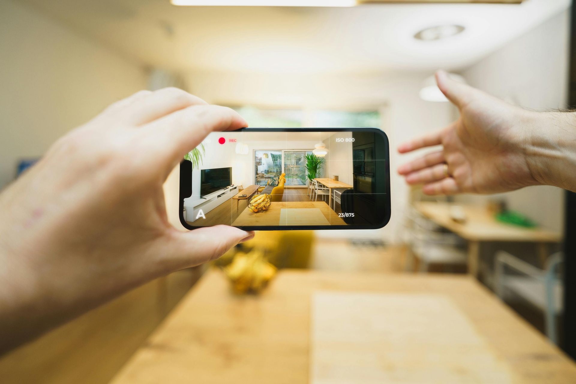 Person holding phone filming an interior space, showing living room with light, wood furniture. Another hand reaches toward the phone.