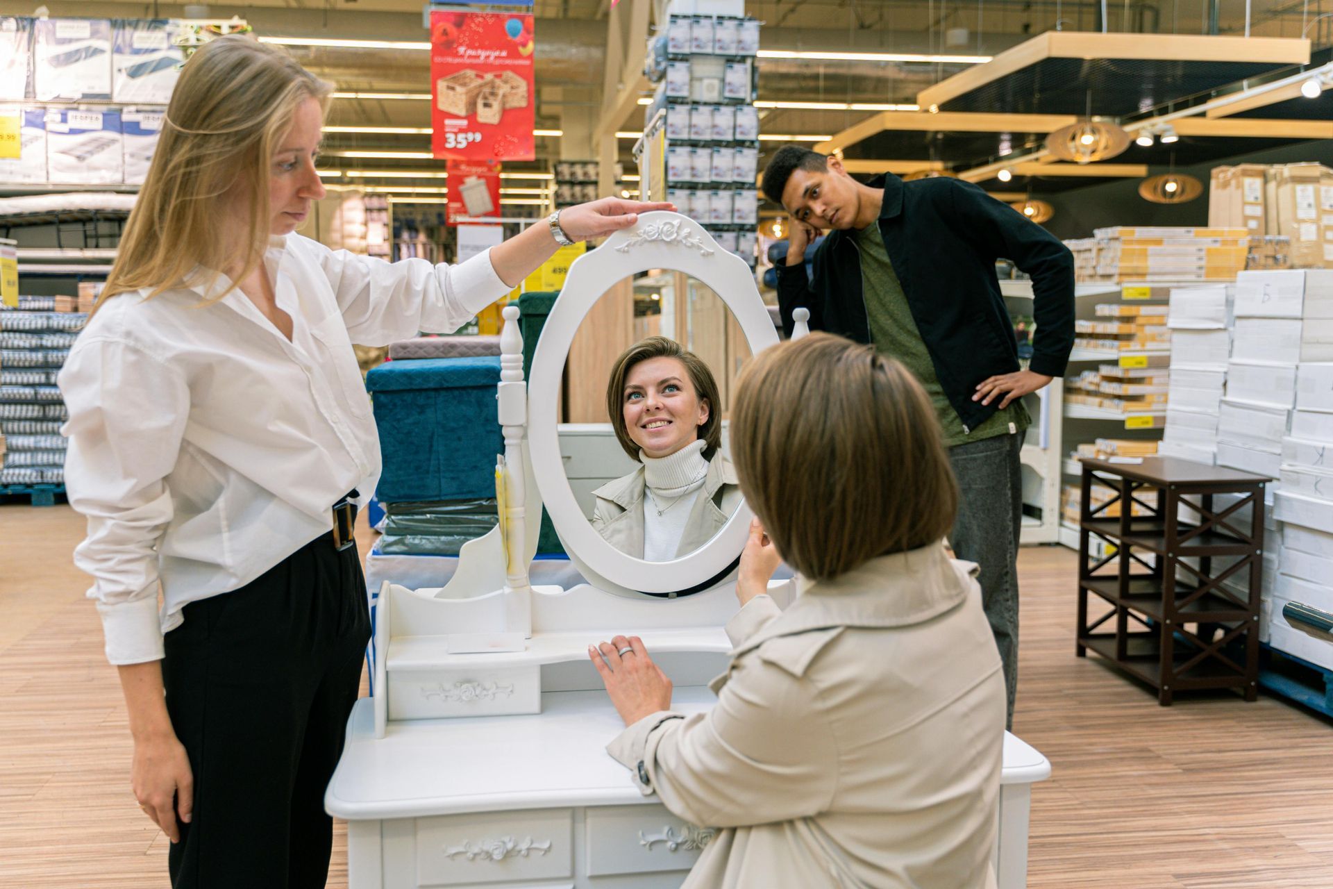 Woman adjusts a mirror for a woman looking in it; a man watches in a store.