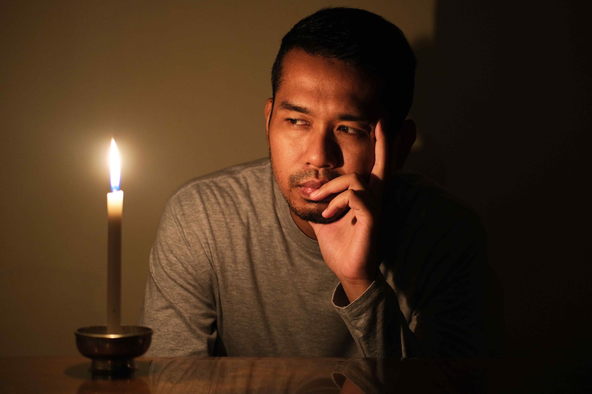 man sitting in the dark staring at a lit candle looking troubled