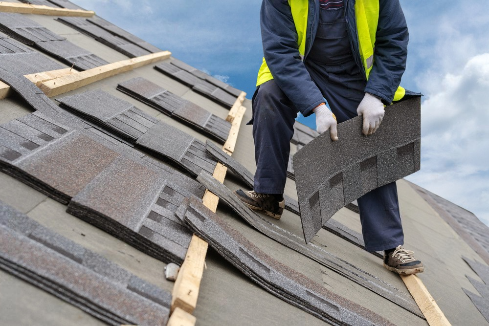 A man is kneeling on a roof holding a piece of shingles.