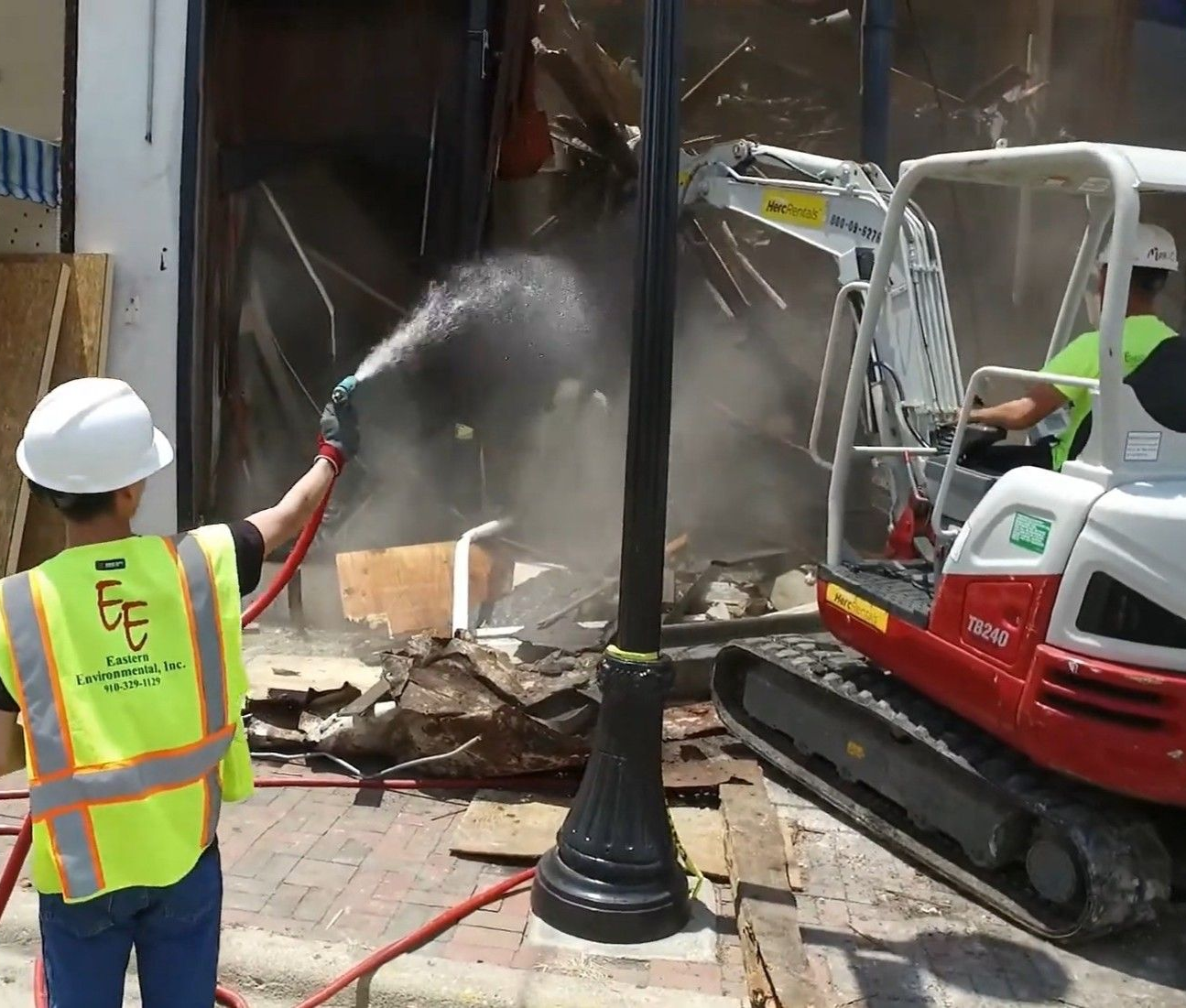 Worker spraying water at debris by a damaged storefront beside a red excavator in Greenville SC