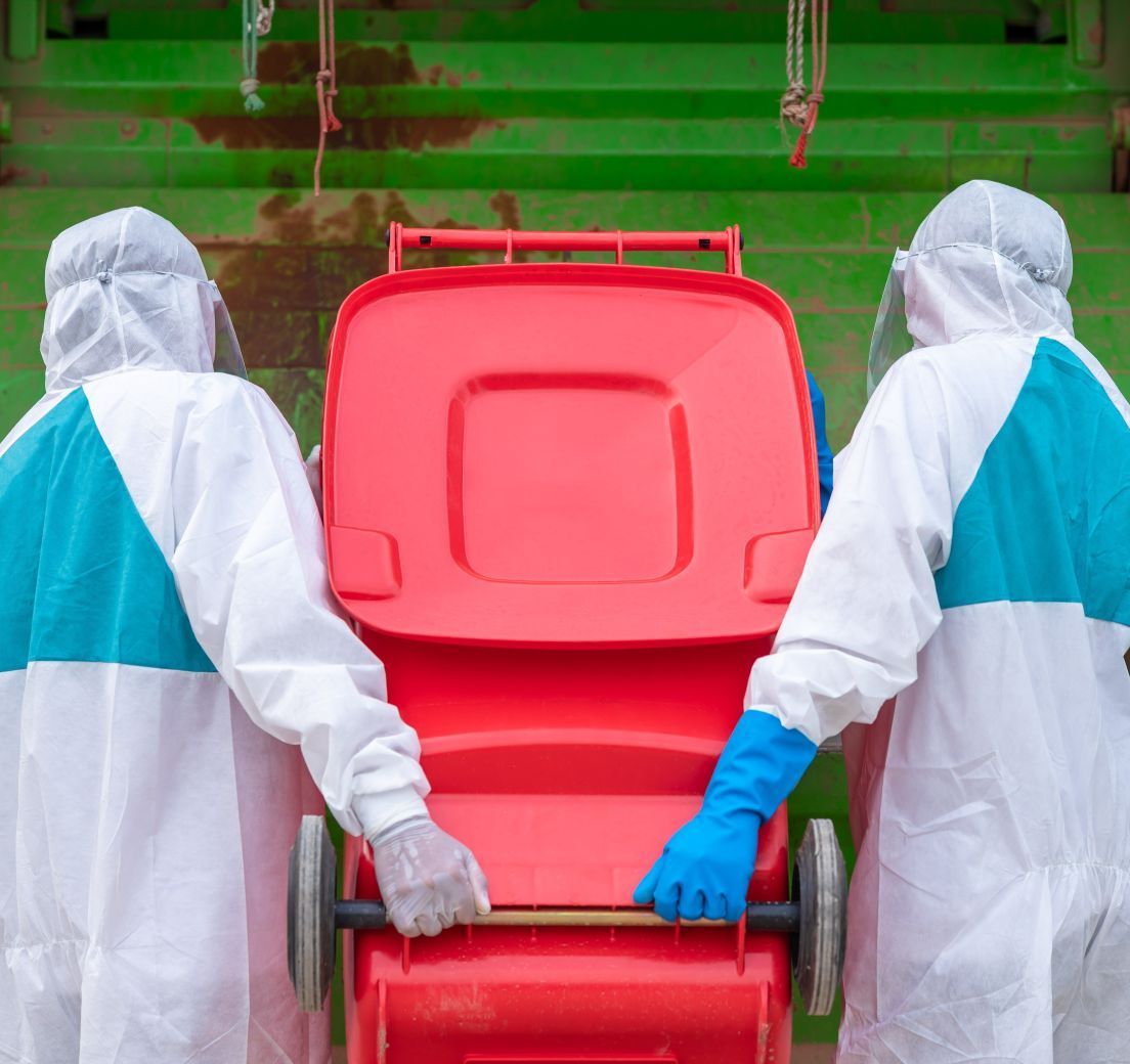 Two people in protective suits are pushing a red trash can.