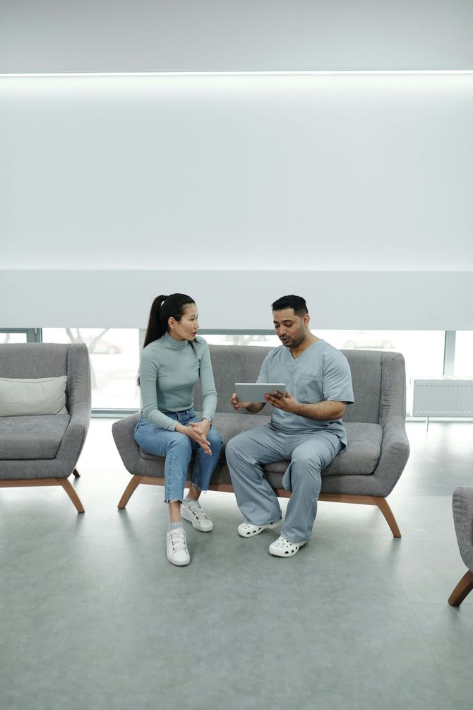 A doctor in scrubs reviews a tablet with a seated patient in a modern waiting room.
