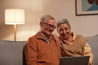Elderly couple smiles while looking at a laptop on a couch in a living room.