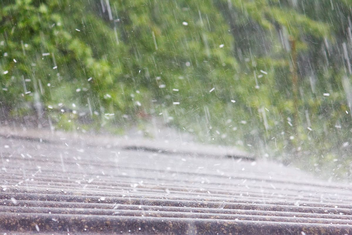 A close up of rain falling on a roof with trees in the background.