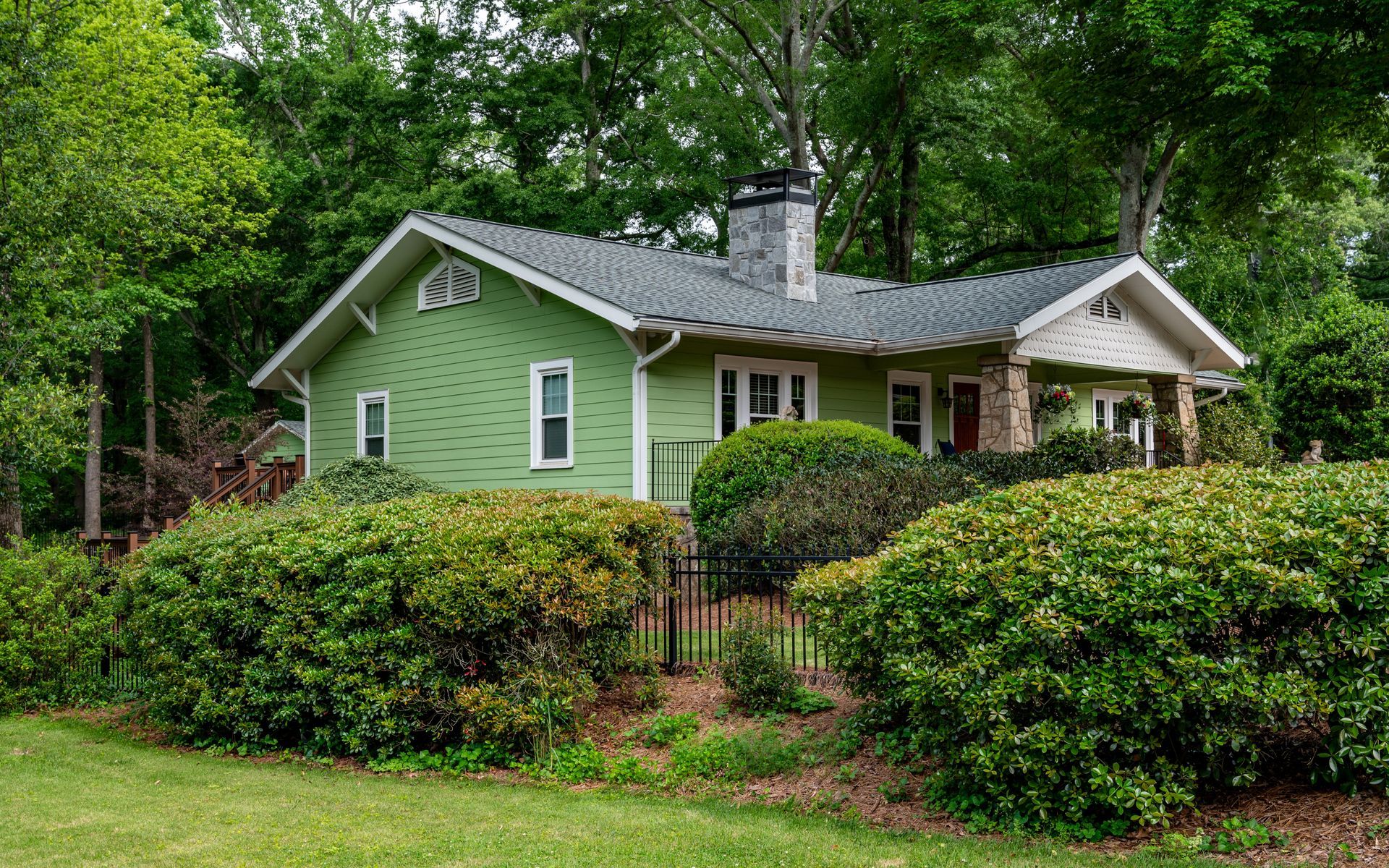A green house with a fence and bushes in front of it is surrounded by trees.