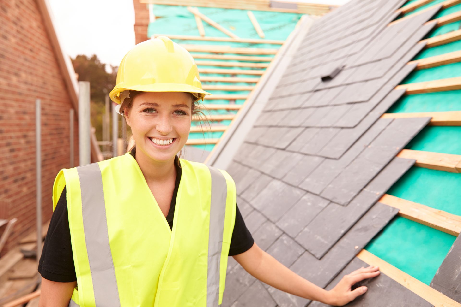 A woman wearing a hard hat and safety vest is standing next to a roof.