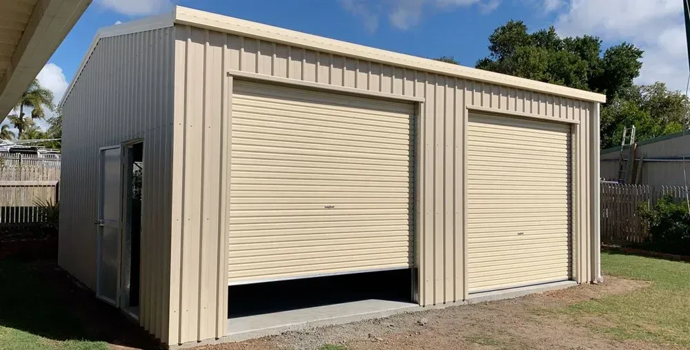 A Beige Two-Bay Metal Shed with Roll-Up Doors and A Side Entry Door — BF Sheds & Garages In Rockhampton, QLD