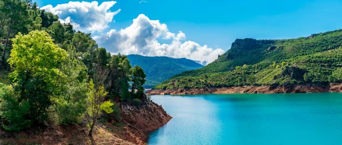 A vibrant blue lake surrounded by lush green mountains and trees under a clear, cloudy sky.