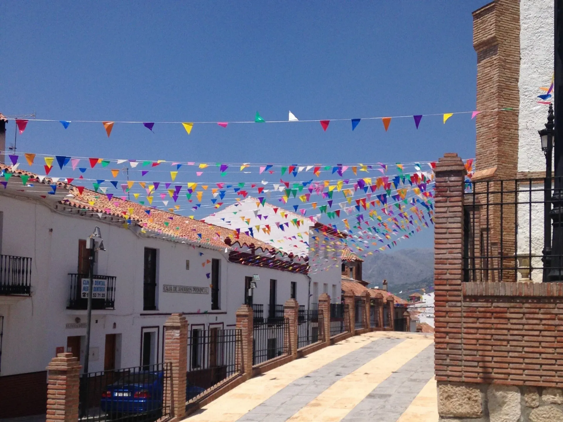 A street in a Spanish town with white buildings, colorful bunting, and a brick tower under a blue sky.