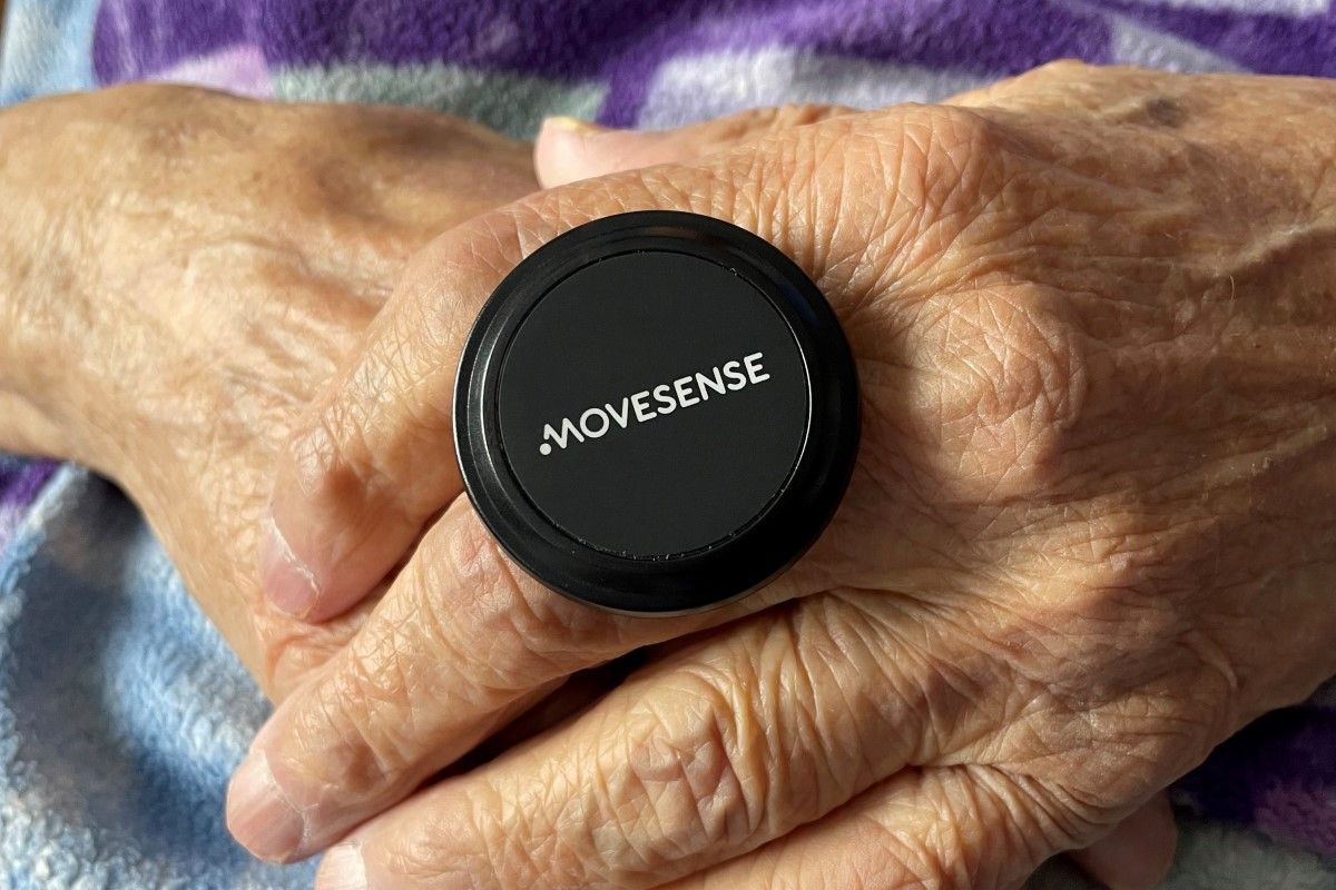 Black Movesense sensor on elderly person's hands. Wrinkled skin and blue fabric background.