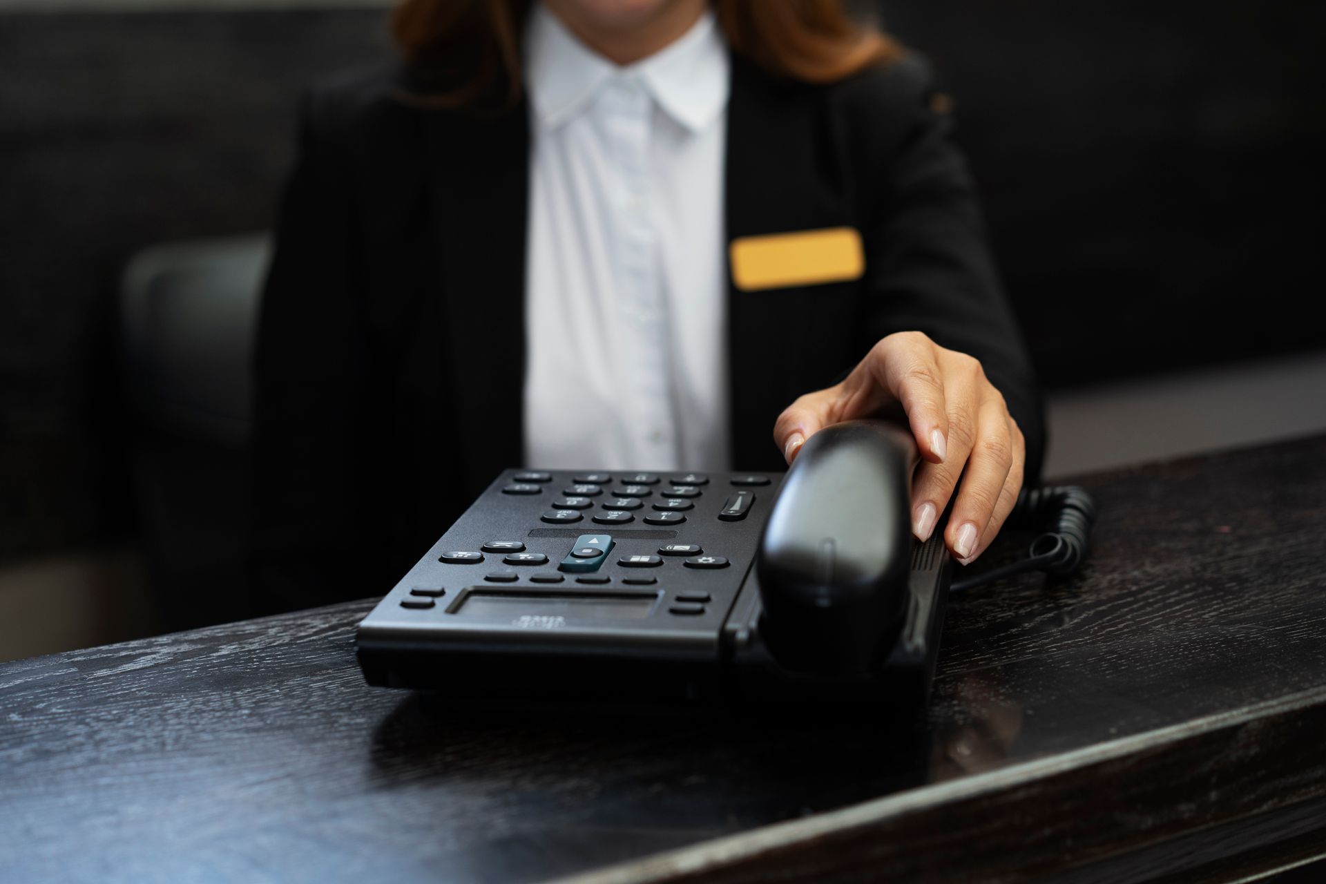 Person in a blazer on a desk preparing to answer a black telephone at a reception desk.