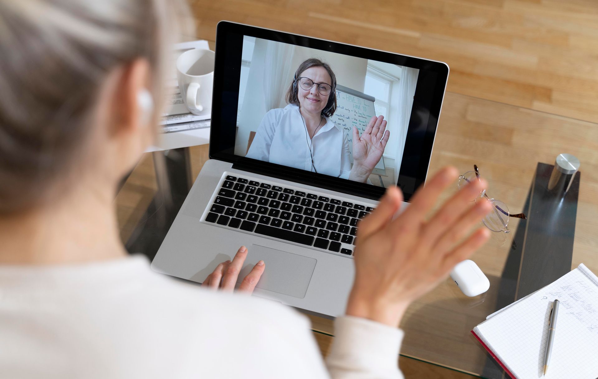 Person in front of laptop waving; another person on the screen waves back, home setting.