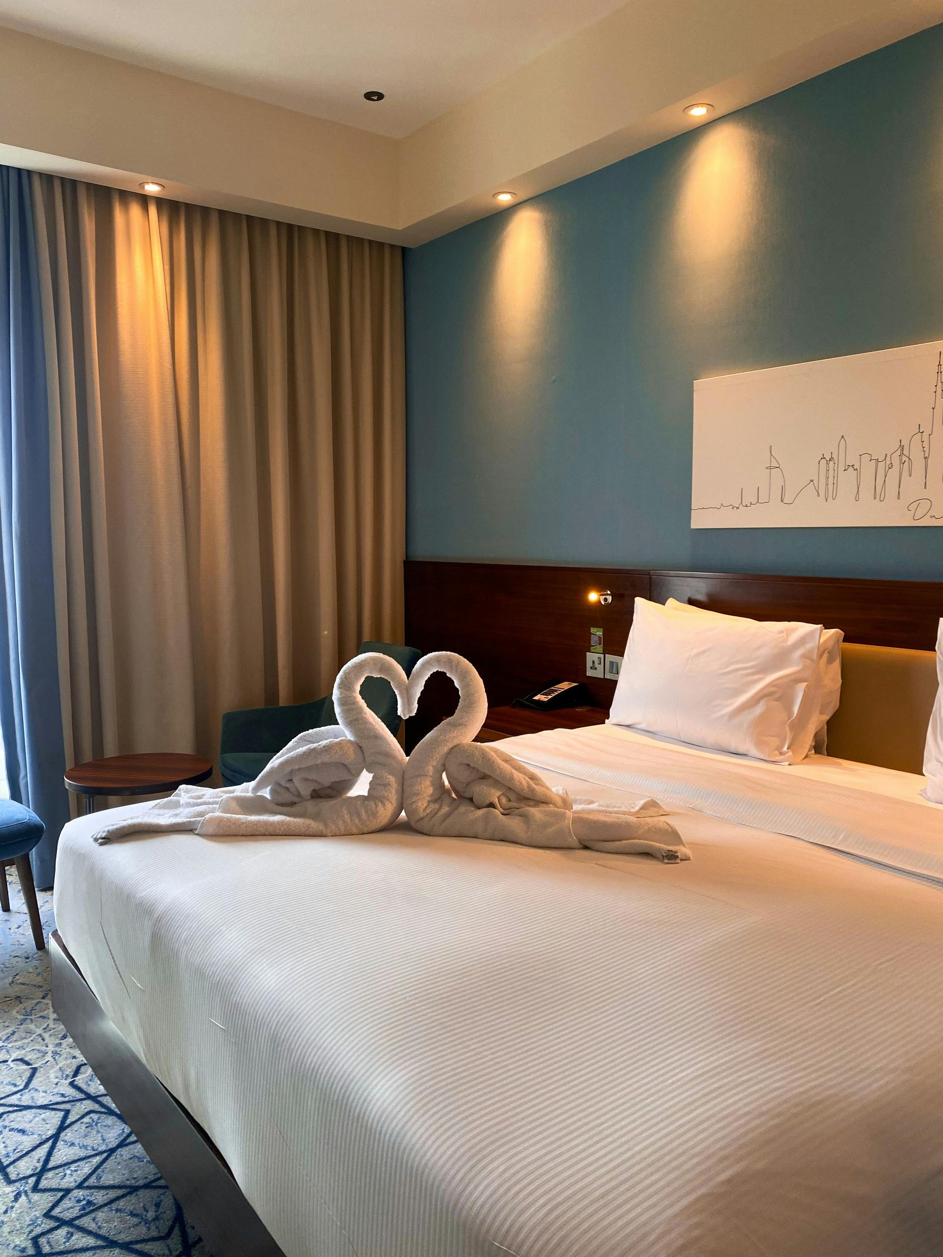 Hotel bedroom with white bed and towel swans, blue wall, brown curtains.