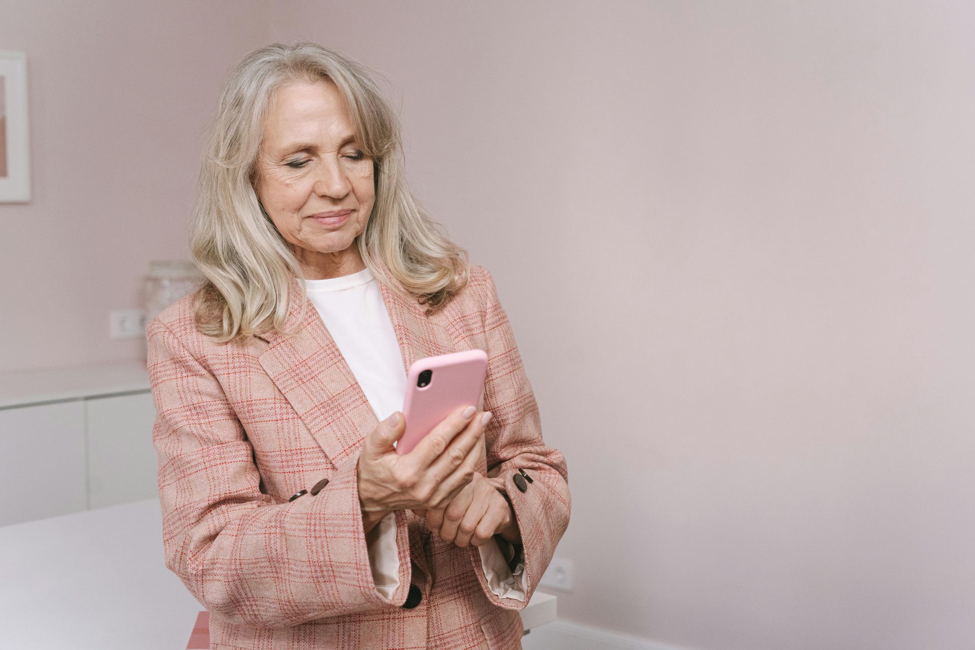 Woman with gray hair in a pink blazer looks at a pink smartphone in a room with a pink wall.