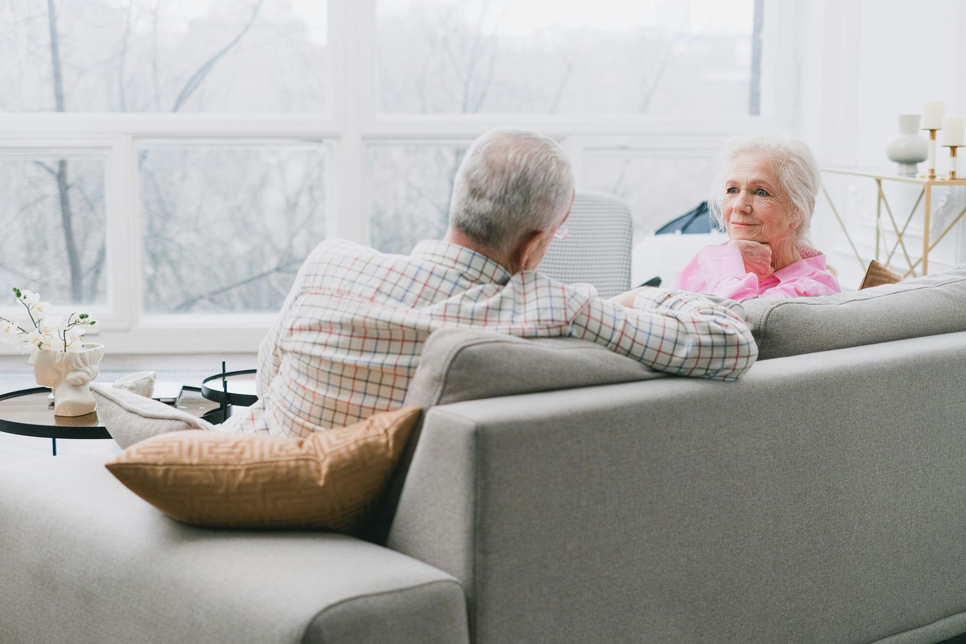 Couple on a gray sofa in a living room, man facing woman, light coming from the window.