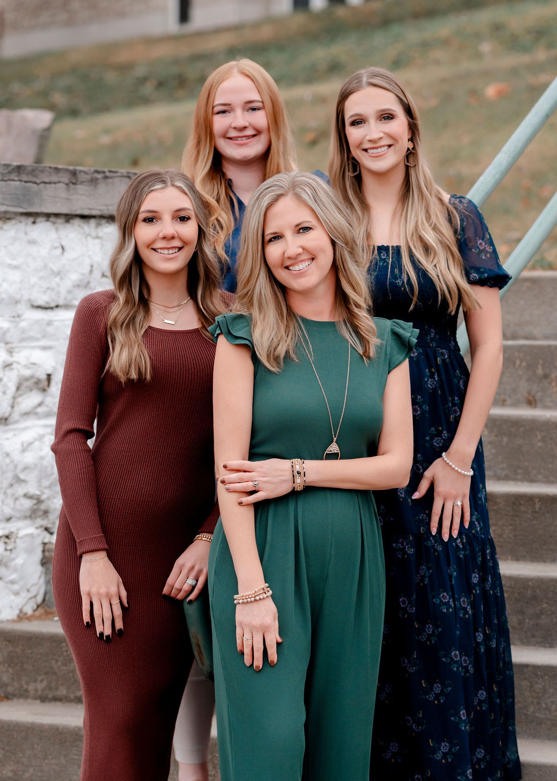 Four women posing outdoors, leaning on a dark metal structure. They wear various business casual outfits, smiling.