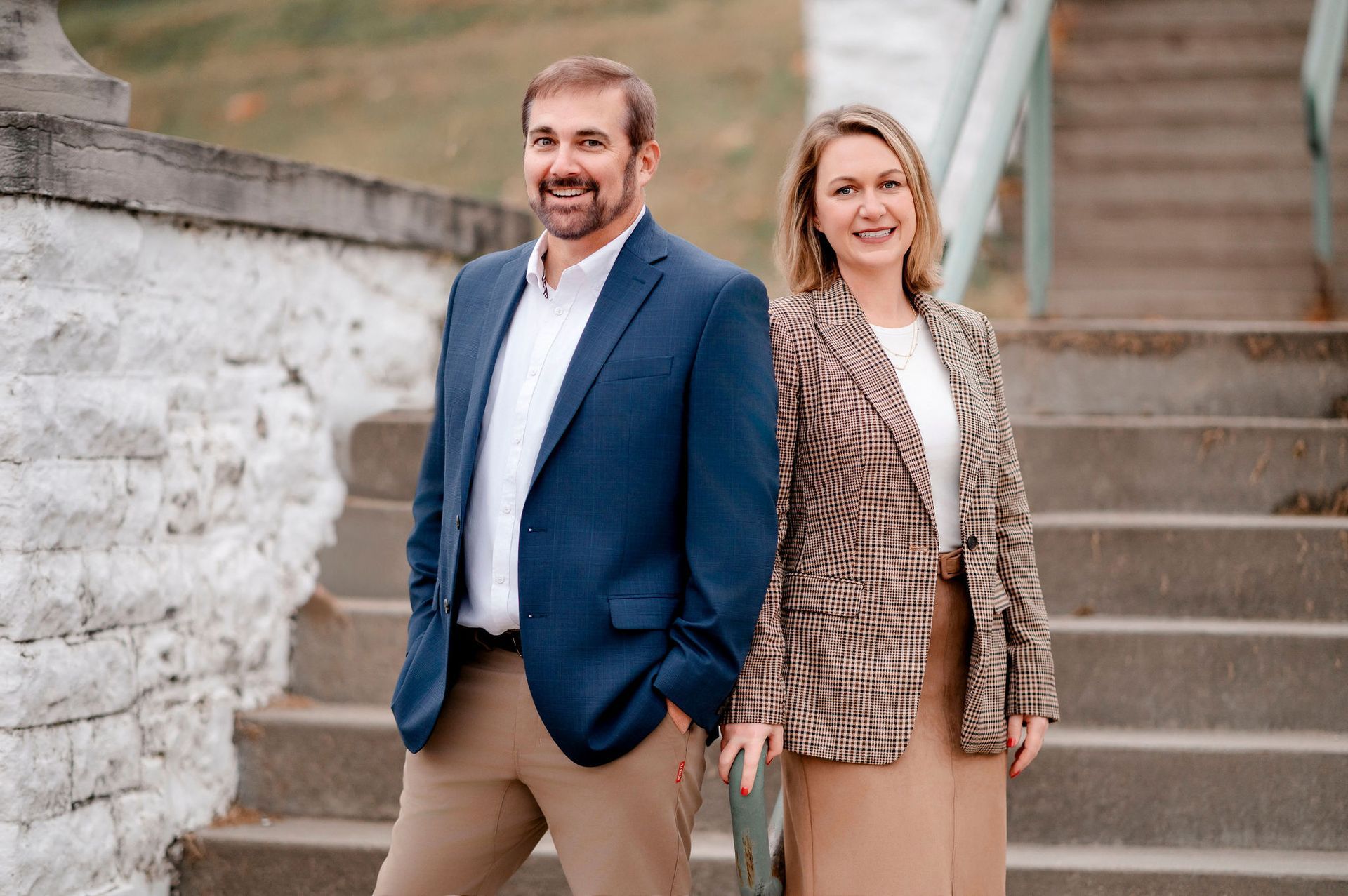 Man in blue blazer and woman in red dress smile outside brick building.