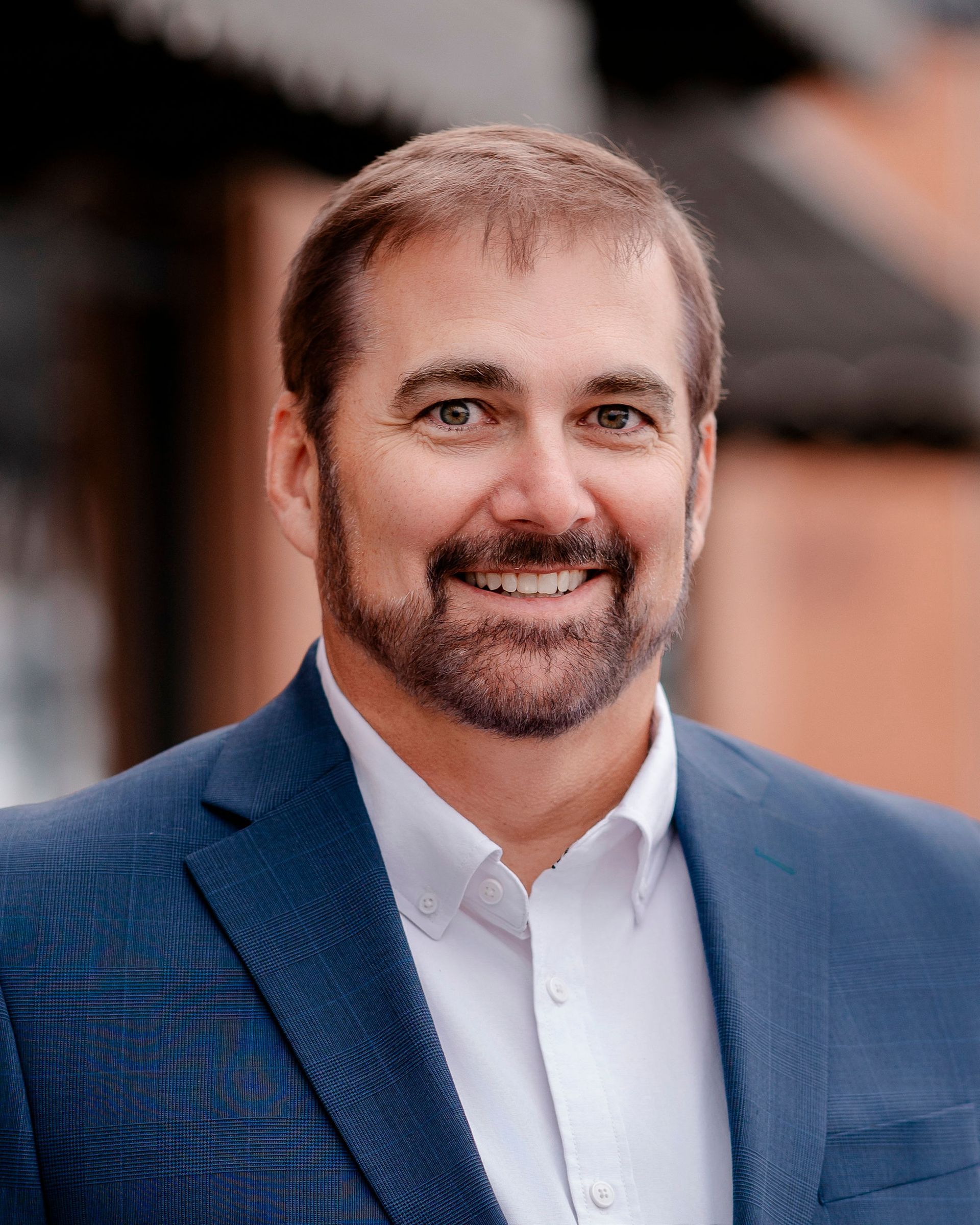 Man with glasses smiles, wearing a dark sweater outdoors.