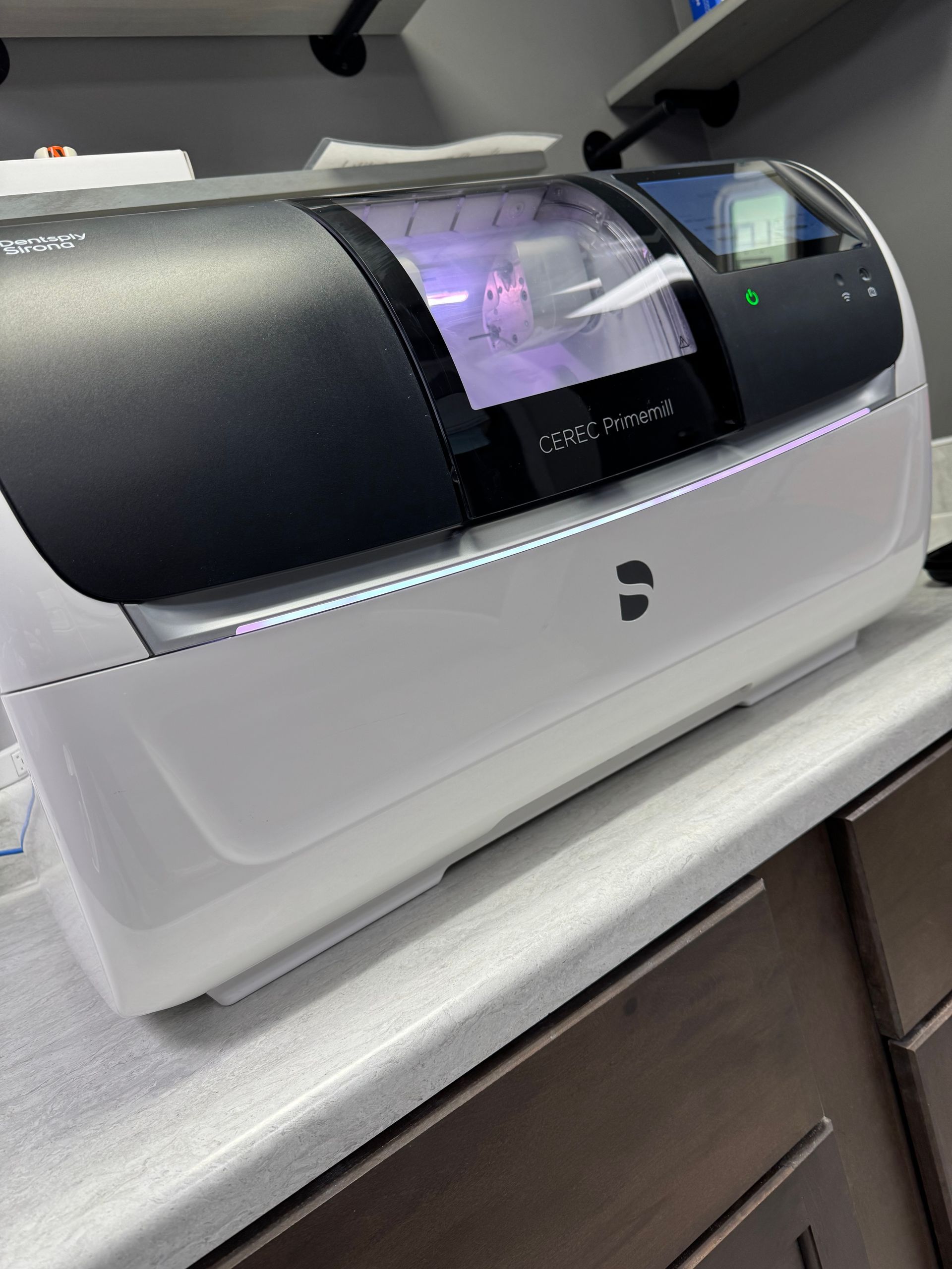 Dental milling machine on a countertop. White and black, with a screen and a visible cutting process.