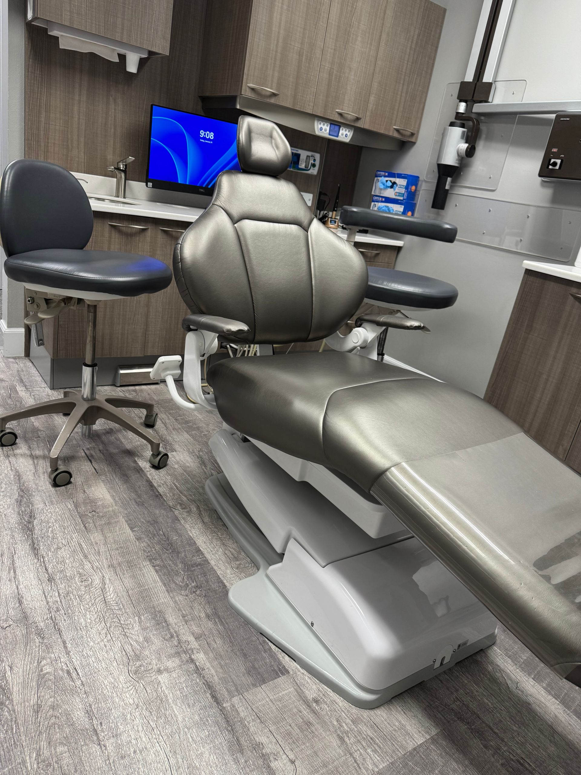 Dental examination room with a patient chair, stool, and cabinets. Gray and blue tones.