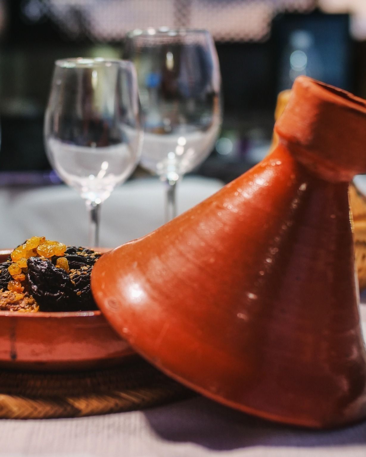 A clay pot filled with food sits on a table next to two wine glasses.