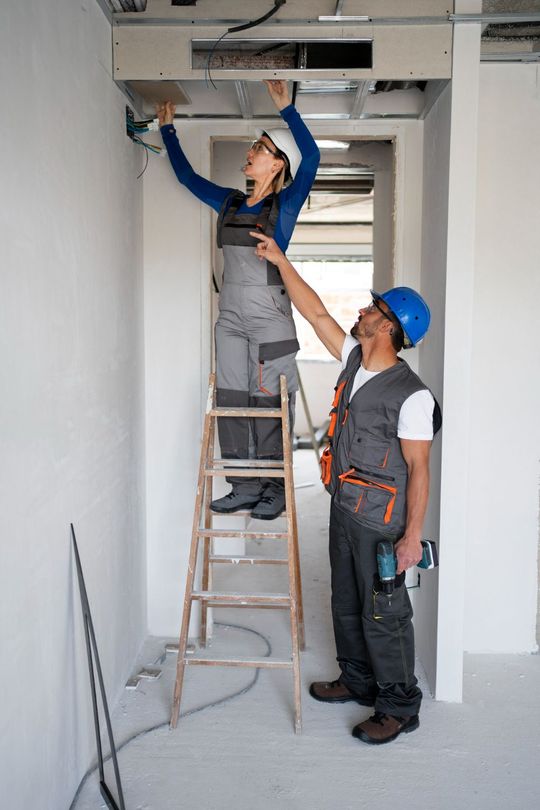 Two construction workers in a doorway: one on a ladder working on ceiling wiring, the other pointing.