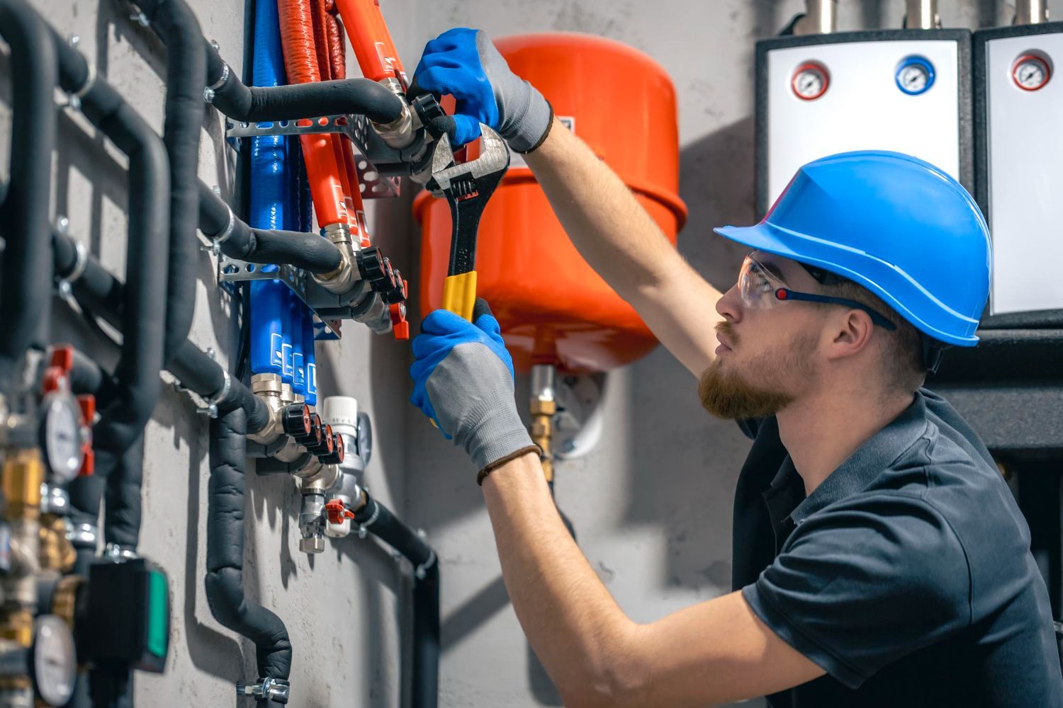 Plumber in blue hat and gloves uses wrench on pipes near red tank.