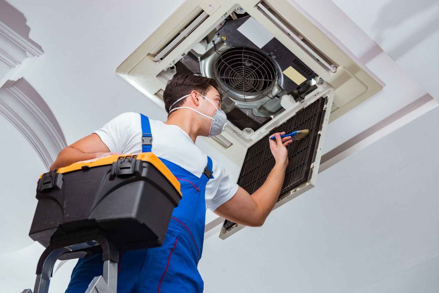 HVAC technician in blue overalls on ladder, servicing ceiling air conditioner; tool box nearby.