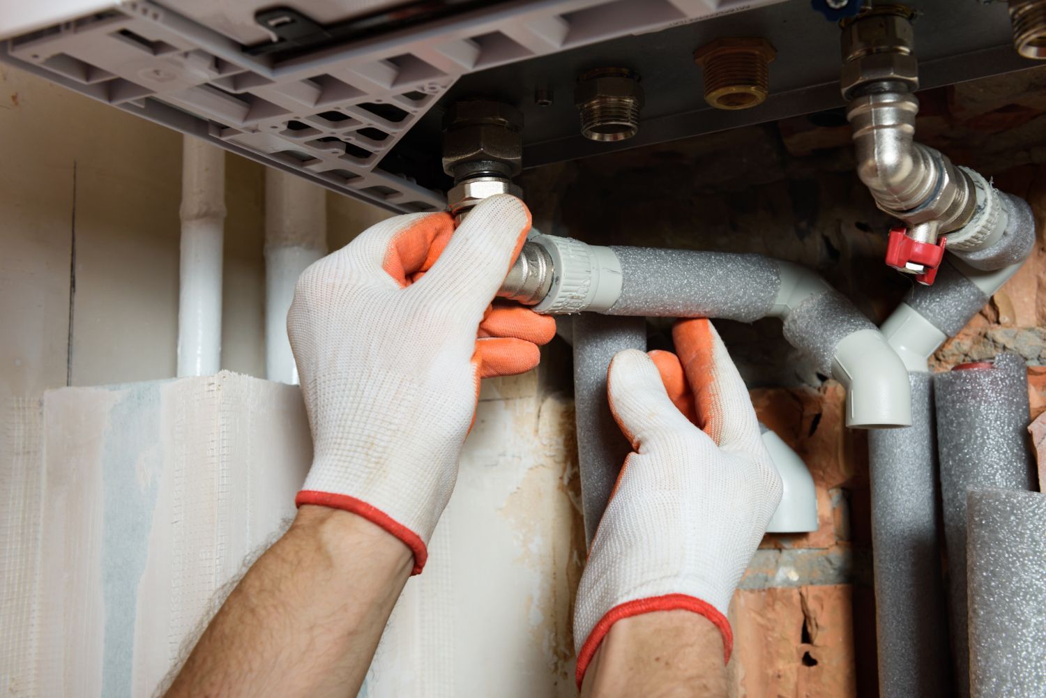 Hands in gloves connecting pipes near a boiler in a brick-walled setting.
