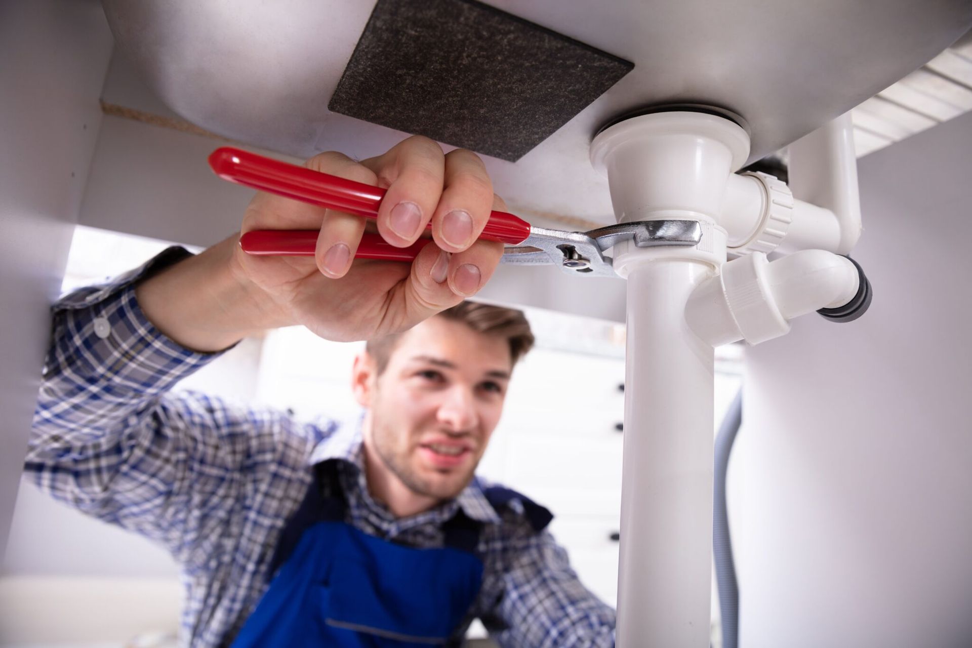 Plumber using a wrench under a sink. White pipe, red handled wrench, and the plumber is wearing blue overalls.