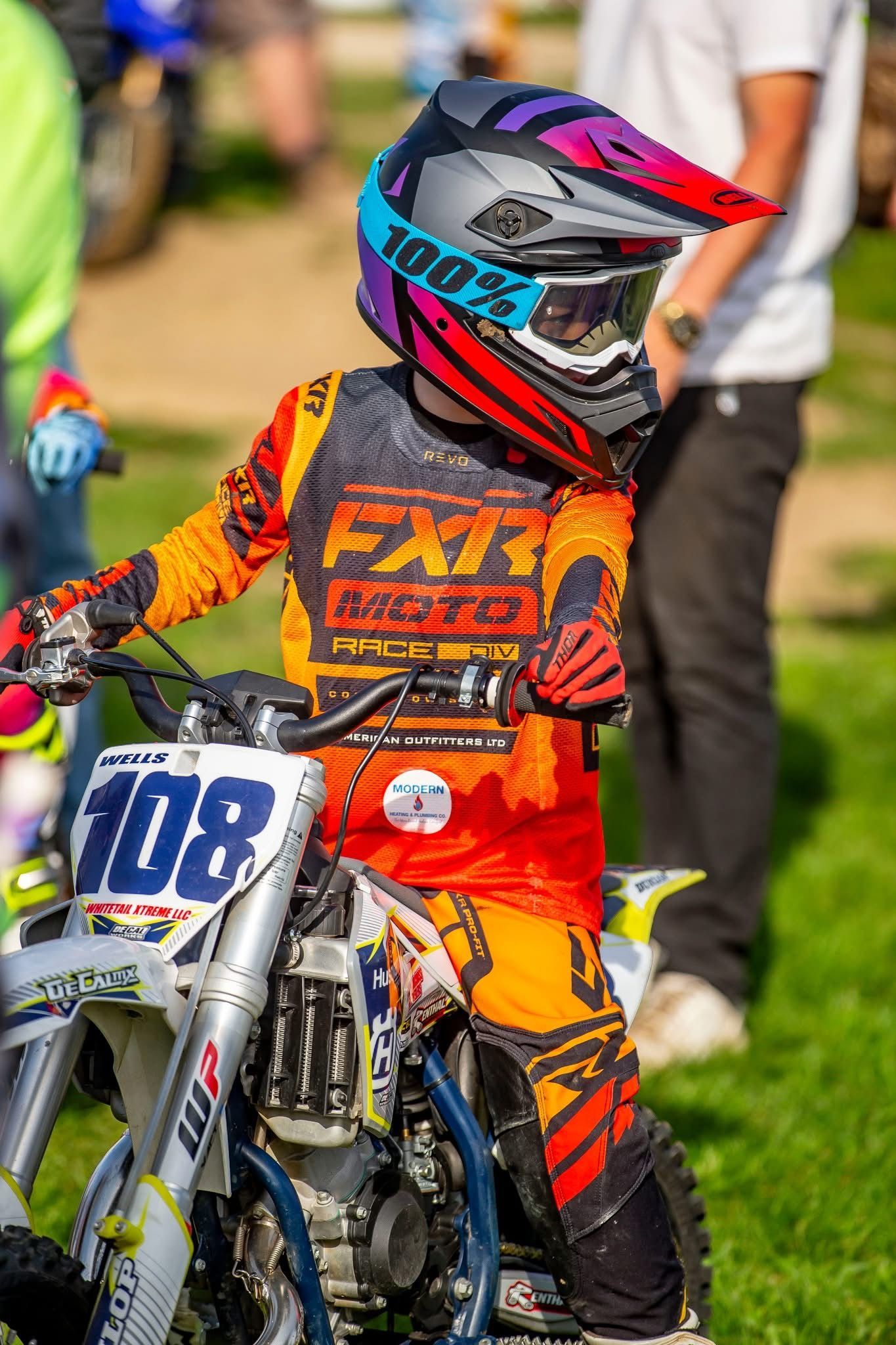 Young rider on a motocross bike, wearing orange and black gear and a colorful helmet, outdoors on a grassy track.