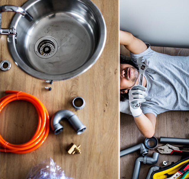 Plumber lying on floor repairing kitchen sink, surrounded by tools and pipes.