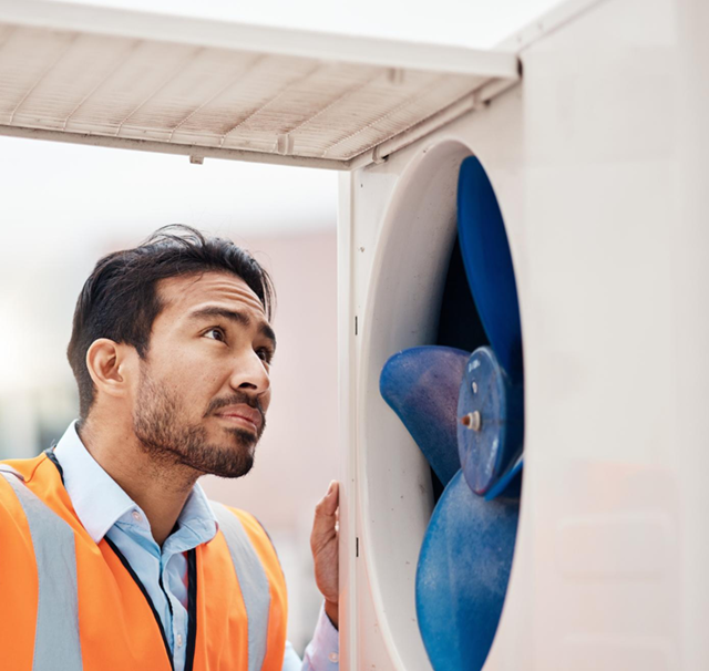 Man in safety vest inspecting large fan inside a white machine, looking up with a thoughtful expression.