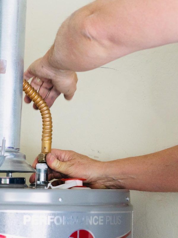 Person connecting a corrugated copper gas line to a water heater. Hands are visible; interior setting.