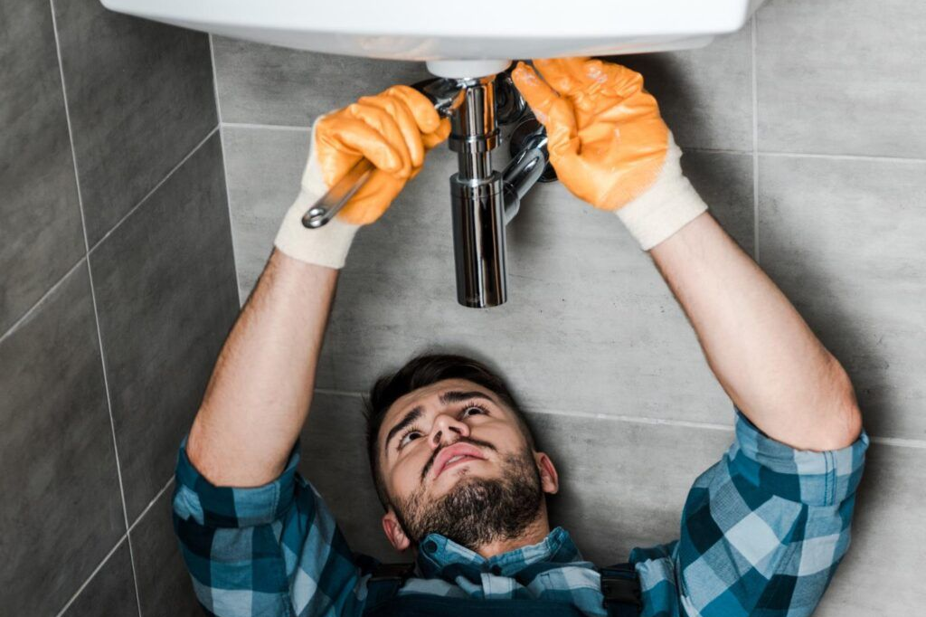 Plumber in orange gloves fixing a sink's pipes with a wrench, wearing a blue plaid shirt, in a bathroom.